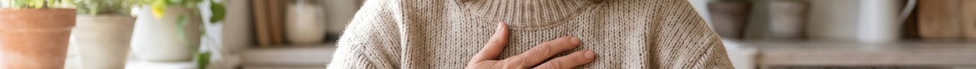 A calm middle-aged woman in a cozy sweater resting her hand over her heart at a sunlit kitchen table with a glass of water and a small dish of spirulina tablets