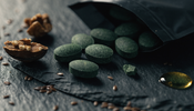 A macro close-up of green spirulina tablets spilling from a matte black pouch onto slate beside flax seeds and walnuts