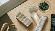 A weekly pill organizer on a light wood table beside a glass of water and a small glass dish of green algae tablets