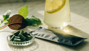A small glass dish of green spirulina tablets on a marble counter beside a glass of lemon water and fresh leafy greens
