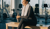 A fully clothed middle-aged woman in long-sleeve athletic wear rests on a gym bench beside a water bottle and a small glass dish of green algae tablets