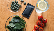 Overhead flat lay on a warm oak table with a glass dish of green algae tablets, a matte black pouch, a bowl of leafy greens, cherry tomatoes and a glass of water with lemon