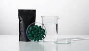 A clean white lab bench holding a glass dish of dark green algae tablets, a matte black supplement pouch, a glass beaker of water, and a petri dish under bright cool lighting