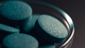 Macro close-up of deep blue-green spirulina tablets piled in a small glass dish against a dark charcoal background