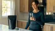 A middle-aged woman in long-sleeve activewear stands in a bright kitchen holding a glass of water, with a matte black supplement pouch and a dish of green algae tablets on the counter.