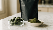 A glass dish of deep green chlorella tablets beside a small bowl of loose green chlorella powder on a light kitchen counter.