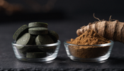 Two small glass dishes side by side on dark slate, one holding green spirulina tablets and the other holding tan ashwagandha root powder with a dried root laid beside it.