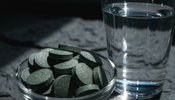 A macro close-up of deep green algae tablets piled in a small glass dish beside a tall glass of clear water on a dark stone surface