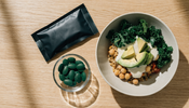 A plant-based grain bowl on a light oak table beside a small glass dish of green algae tablets and a matte black supplement pouch
