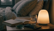 An evening bedside table with a warm lamp, an open book, a glass of water and a small dish of green chlorella tablets