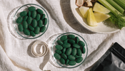 An overhead flat lay on linen with two dishes of green algae tablets, a coiled measuring tape and a bowl of fresh vegetables