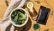 Overhead flat lay on an oak table with a bowl of leafy greens, a glass of water with lemon, a plain matte black supplement pouch, and a small glass dish of green algae tablets