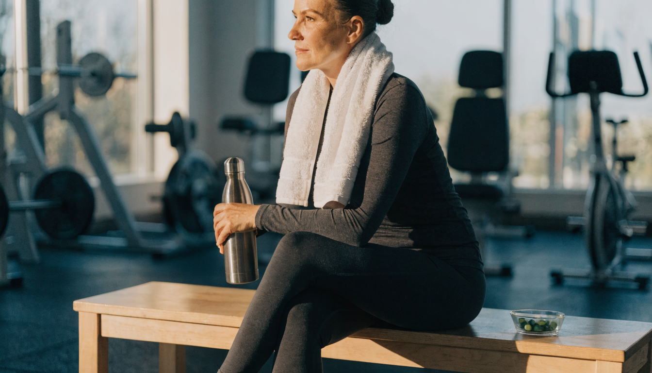 A fully clothed middle-aged woman in long-sleeve athletic wear rests on a gym bench beside a water bottle and a small glass dish of green algae tablets