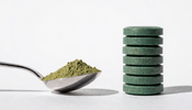 A measuring teaspoon of green spirulina powder beside a neat stack of round green spirulina tablets on a white surface