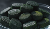 Extreme macro photograph of dark green algae tablets resting in a small glass dish, lit from the side against a dark moody background