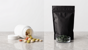 An unlabeled white supplement bottle with colorful tablets beside a matte black pouch and a small glass dish of green algae tablets on a light grey stone counter