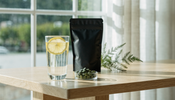 A tall glass of water with a lemon slice beside a plain matte black supplement pouch and a small glass dish of loose green algae tablets on a pale oak table in bright daylight