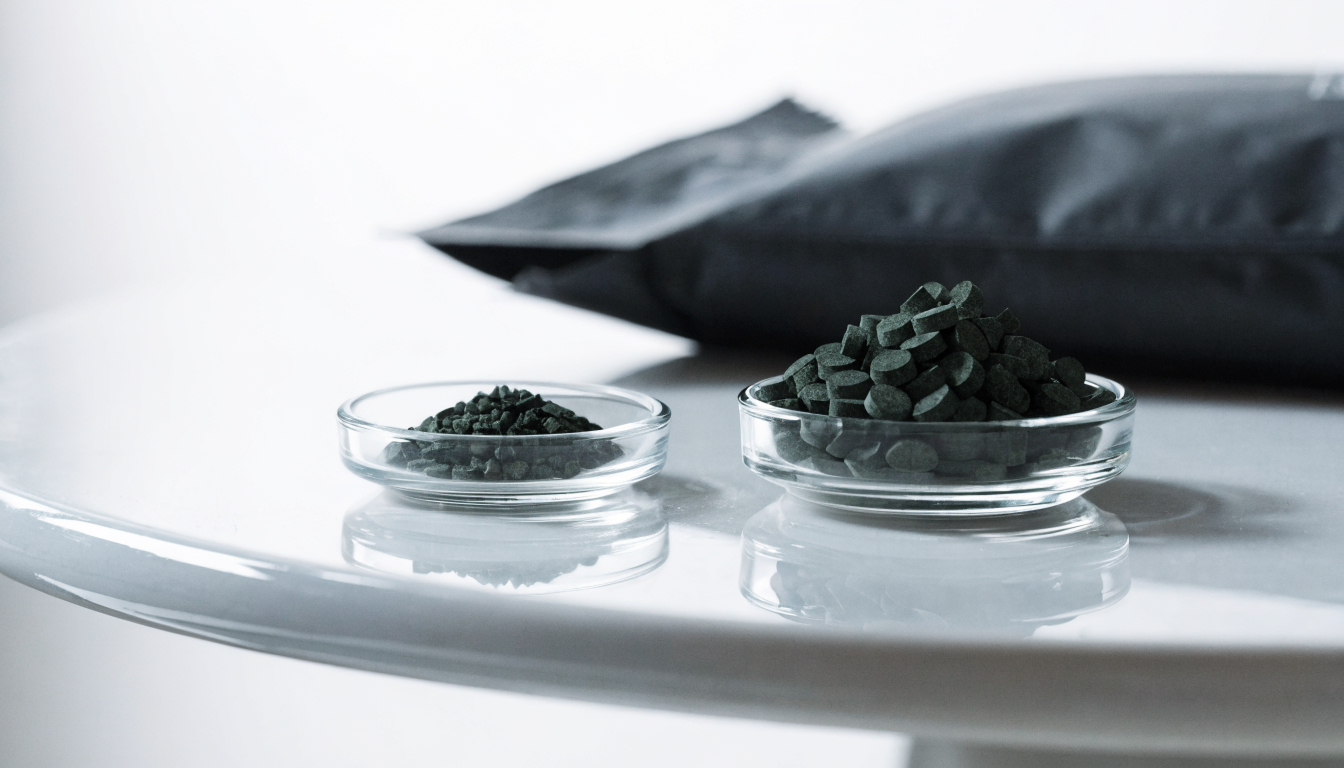 Two small clear glass dishes side by side on a white surface, one holding a small measured portion of dark green algae tablets and the other a much larger heaped portion, with a plain matte black supplement pouch behind them.