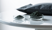 Two small clear glass dishes side by side on a white surface, one holding a small measured portion of dark green algae tablets and the other a much larger heaped portion, with a plain matte black supplement pouch behind them.