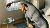A middle-aged woman in long-sleeved grey activewear doing a gentle standing stretch in a sunlit living room, with a glass of water and a small dish of green algae tablets on a side table.