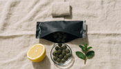 Overhead flat lay on natural linen of an opened plain matte black supplement pouch with dark green algae tablets spilling into a small glass dish, beside a halved lemon and a sprig of mint.