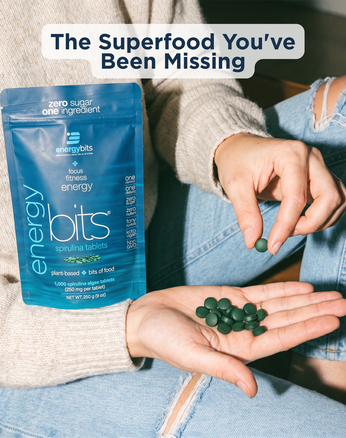 A person in a beige sweater and ripped blue jeans holds a blue pouch of Energy Bits supplement next to their open palm, which contains dark green tablets. A text overlay at the top reads 'The Superfood You've Been Missing'.