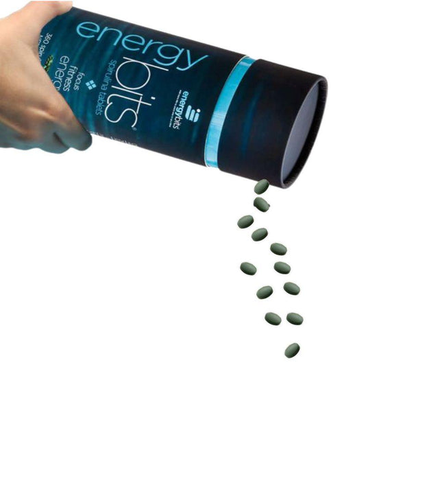 Hand pouring small spirulina tablets from an ENERGYbits® container against a white background.