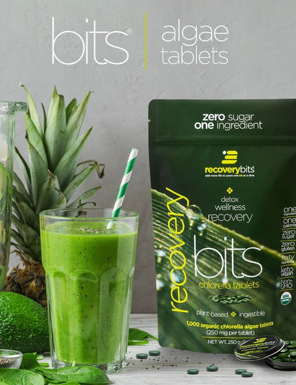 A bag of RECOVERYbits® chlorella algae tablets next to a green smoothie, spinach leaves, an avocado, and a pineapple